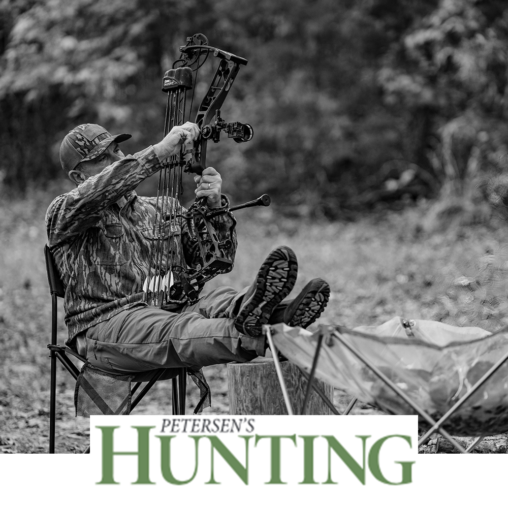Meindl Comfort Fit Camp Moc Slip-On – As Featured in Petersen's Hunting Magazine – Leather Moccasin Camp Shoe Worn by Bowhunter in Camouflage Inspecting Compound Bow at Campsite With Fire Visible