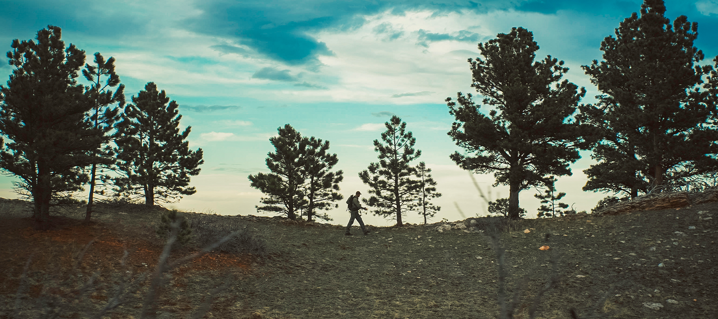 Lone Hiker Silhouetted Against a Dramatic Sunset Sky Walking Across a Rocky High Desert Ridge Between Scattered Pinon Pine Trees