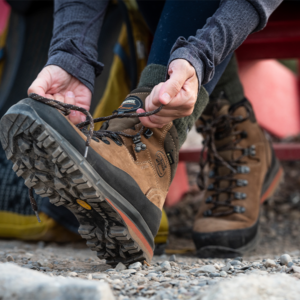Meindl Lady Vakuum Hunter – Brown Suede Gore-Tex Women's Hiking Boot Being Laced Up at a Trailhead With a Second Boot and Yellow Backpack Visible in Background