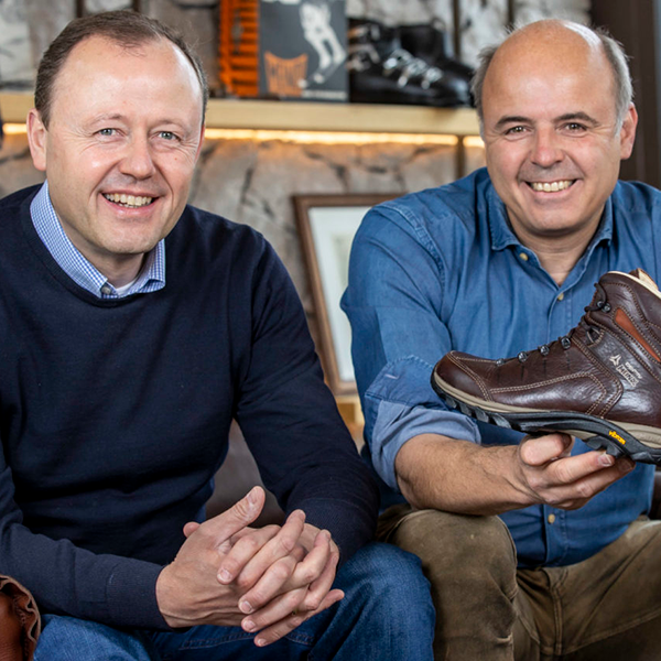 Meindl Brand History – Two Smiling Men Seated Together in a Meindl Showroom Holding a Brown Leather Hiking Boot Surrounded by Vintage Meindl Memorabilia